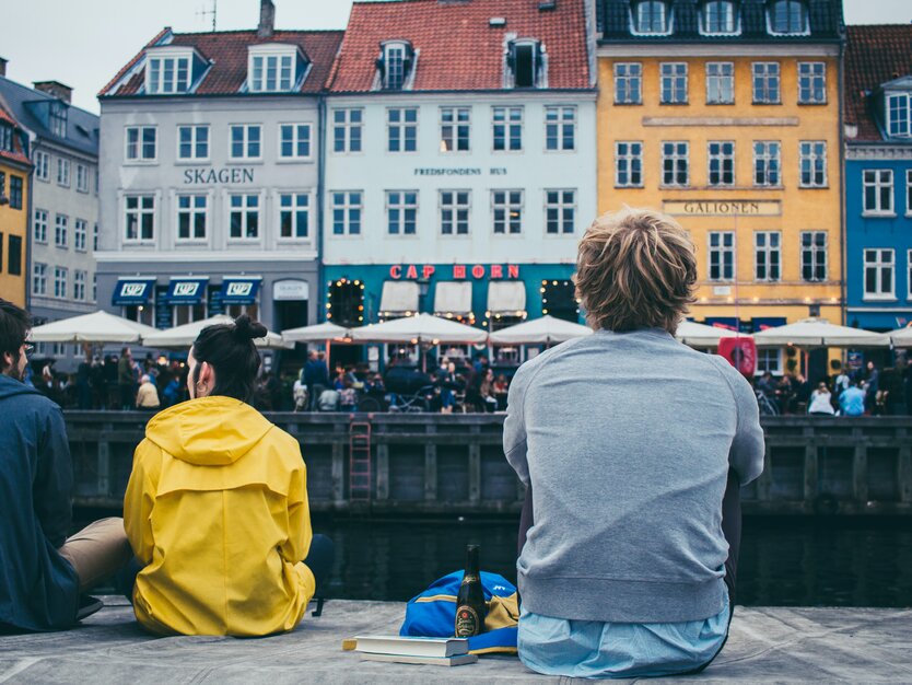 Städtereise Kopenhagen, Menschen sitzen am Steg des Hafens in Kopenhagen mit Blick auf die Häuser am anderen Ufer