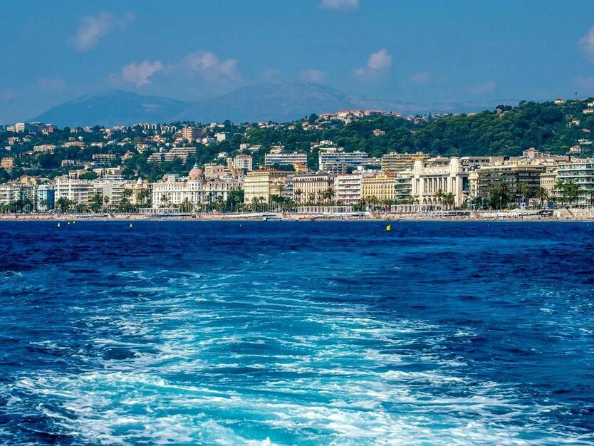 Städtereise Nizza, Frankreich, Bootsfahrt Nizza, Cote d Azur, Blick über die Wellen des Bootes auf die Promenade, Häuser und Bäume an der Küste, Gebirge im Hintergrund, gutes Wetter